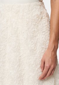 Jupe blanche à fleurs présentant des appliqués floraux texturés en couches. Un gros plan montre une main touchant délicatement le tissu, mettant en valeur les détails et la texture.