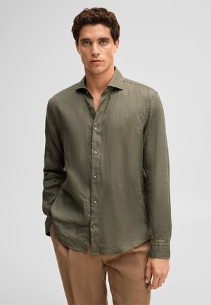 Homme portant une chemise boutonnée à manches longues vert olive et un pantalon brun clair, debout devant un fond gris clair uni.