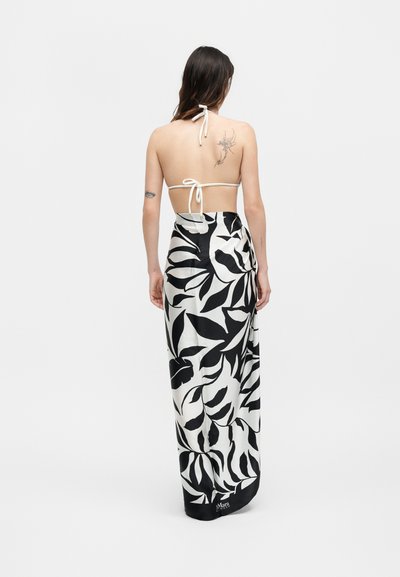 Γυναίκα με μακριά σκούρα μαλλιά που φοράει λευκό τοπ με τιραντέλα δεμένο πίσω και μακριά φούστα με ασπρόμαυρα φύλλα, γυρισμένη από την πλάτη.