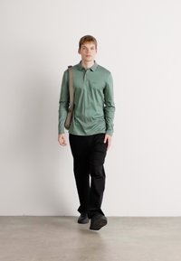 Light green long-sleeve polo shirt with a collar, paired with black pants and dark shoes, worn by a person standing against a neutral background.