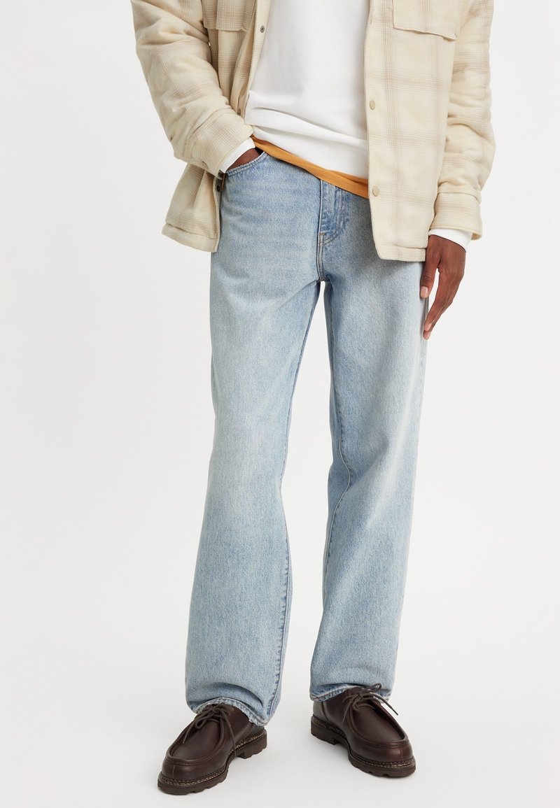 Homme portant un jean denim bleu clair, une veste à carreaux beige, des chemises blanches et orange superposées, et des chaussures marron foncé à lacets, debout avec une main dans la poche.