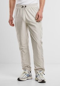 Lichtbeige, lichte joggers met een elastische taille en trekkoord. Heeft witte zijpijpen, een gladde textuur en een taps toelopend ontwerp.