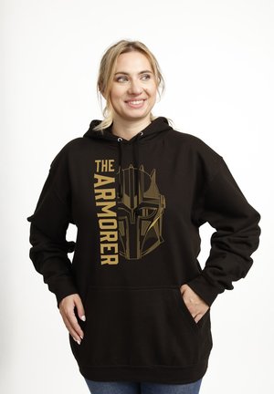 Mujer sonriente con cabello rubio que lleva una sudadera negra con un casco Mandaloriano dorado y el texto "THE ARMORER."