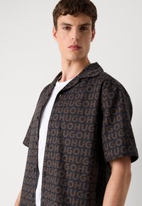 Camisa de manga corta en tejido oscuro con un patrón repetido del logo "Hugo" en color marrón, que cuenta con un cuello puntiagudo y cierre de botones.