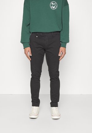 Person trägt dunkle Slim-Fit-Hosen, weiße Sneaker und einen grünen Langarm-Sweatshirt mit kleinem weißem Berglogo auf der Brust.