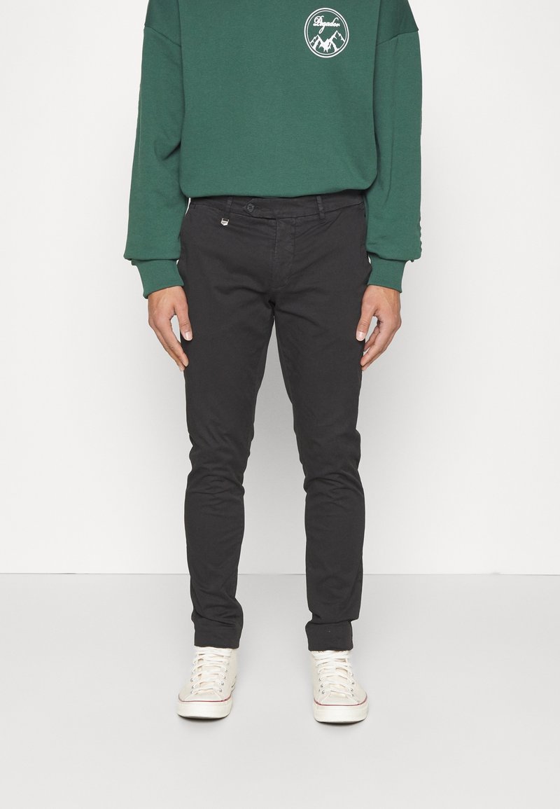 Personne portant un pantalon slim foncé, des baskets blanches et un sweat-shirt vert à manches longues avec un petit logo de montagne blanc sur la poitrine.