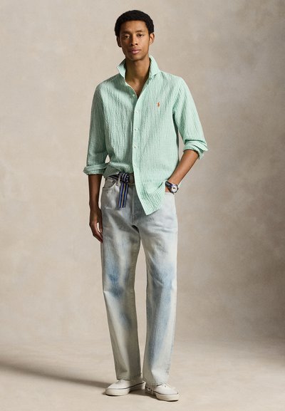 Fiatal férfi világoszöld texturált ingben, világoskék farmerrel, csíkos övvel, fehér tornacipővel, bézs háttér előtt áll.