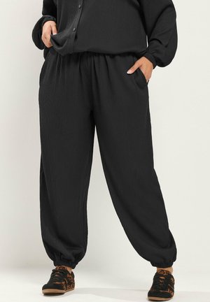 Vrouw draagt losse zwarte textuurbroeken met elastische enkels, een bijpassende zwarte top en zwarte sneakers met bruine luipaardprint details.