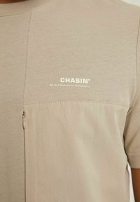 Chemise beige à manches courtes avec une texture lisse, dotée d'une poche zippée et d'un logo embossé "CHASIN'" sur le devant.