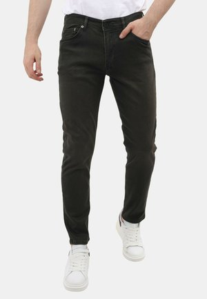 Man draagt strakke zwarte jeans en witte sneakers, met één hand in de zak en een wit overhemd ingestopt.