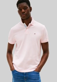 Polo rose clair avec un col, arborant une rayure sous le col et un petit logo sur la poitrine ; manches courtes.