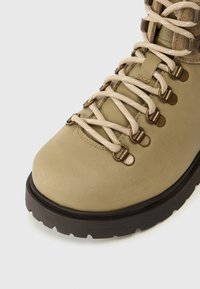 Botte de randonnée en cuir beige avec des lacets beiges et œillets en métal. Elle possède une semelle en caoutchouc texturé et un design à bout rond.