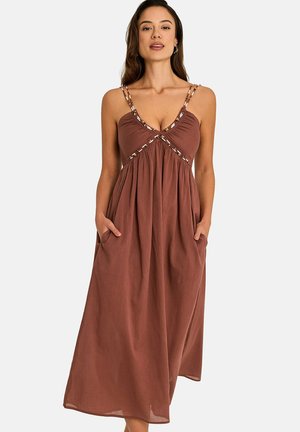 Femme portant une robe marron maxi sans manches avec des bretelles doubles perlées et les mains dans les poches, debout devant un fond uni.