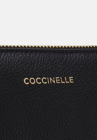 Černá kožená peněženka s texturovaným povrchem, zdobená zlatým zipem a zlatým reliéfním logem "COCCINELLE" na přední straně.