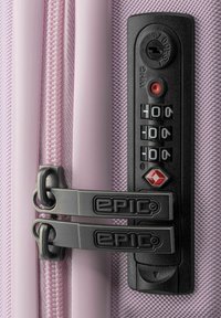 Valigia rigida viola con finitura testurizzata, dotata di doppia zip grigia, lucchetto a combinazione e un emblema di sicurezza rosso.