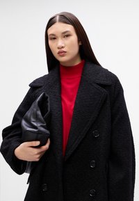 Melns divrindas mētelis ar teksturētu audumu, uzvilkts virs sarkanas turtlenecka blūzes, tur rokās melnu ādas somiņu. Gludas matu frizūras, neitrāls fons.
