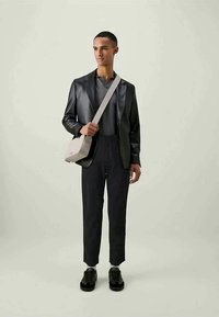 Homem a usar um blazer de couro preto, camisa escura, calças pretas, ténis pretos e uma mala cruzada bege, de pé contra um fundo claro.