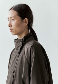 Brązowa, wodoodporna kurtka z wysokim kołnierzem, zapinana na zatrzaski, o gładkiej, fakturowanej powierzchni. Klasyczny, funkcjonalny design.