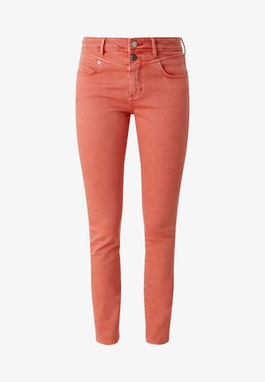 Vrouw die loopt, draagt een losse rode gebreide trui met V-hals, koraal opgerolde broek en witte vetersneakers tegen een neutrale achtergrond.