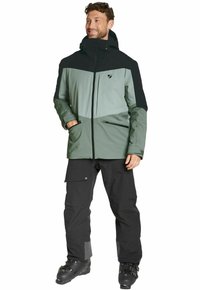 Herren Skijacke in Schwarz und Salbeigrün. Mit Reißverschluss vorne, Kapuze und Reißverschlusstaschen. Kombiniert mit schwarzen Skihosen und Stiefeln.