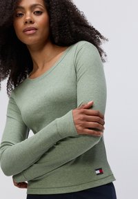 Langsleeve, geribbelde top van lichtgroene stof. Heeft een ronde halslijn en een klein logo-patch aan de onderkant. Zachte textuur.