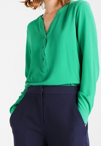 Vihreä pitkähihainen paita, jossa on V-aukko ja kolme nappia edessä, yhdistettynä tummansinisiin korkeavyötäröisiin housuihin. Tasainen kangas.