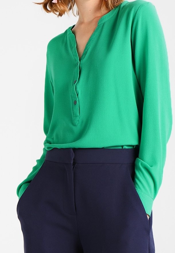 Vihreä pitkähihainen paita, jossa on V-aukko ja kolme nappia edessä, yhdistettynä tummansinisiin korkeavyötäröisiin housuihin. Tasainen kangas.