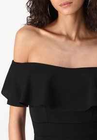 Žena v černých šatech bez ramínek s volánkovým detailem, odhalující odkryté ramena a krk na jednobarevném světlém pozadí.