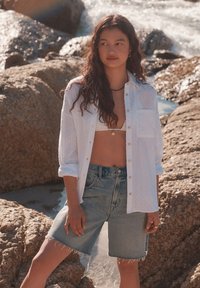 Camicia bianca con bottoni e motivo a trama, indossata sopra una maglia chiara e pantaloncini di jeans a vita alta con bordi sfrangiati. Ambientazione naturale.