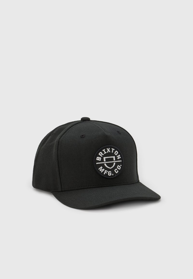 Crna kapa napravljena od čvrstog materijala, s bijelim kružnim logotipom koji sadrži "Brixton MFG Co." Kapa ima strukturirani oblik i ravni vizir.
