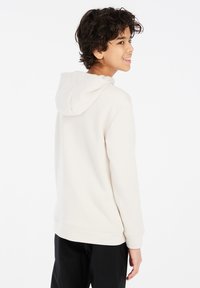 Felpa beige con cappuccio regolabile, polsini e orlo a costine, caratterizzata da una texture morbida. Indossata con pantaloni neri, che mostrano una palette di colori neutra.