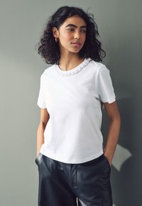 Witte T-shirt met een gestructureerde ronde halslijn; model draagt zwarte leren broek. De stof oogt glad en lichtgewicht.