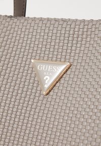 Harmaa kudottu käsilaukku, jossa on kolmion muotoinen kultasävyinen logolevy, jossa lukee "GUESS" ja kysymysmerkki. Rakenteellinen pinta, jossa on hieno kiilto.