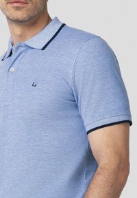 Jasnoniebieska koszulka polo z fakturowanej tkaniny, z dwoma guzikami oraz kontrastowym granatowym wykończeniem na kołnierzyku i rękawach. Emblemat na piersi.