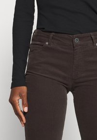 Bruine ribfluwelen broek met een slim fit, voorzien van een knoopsluiting, zijzakken en een zachte textuur. Een zwart shirt met lange mouwen is gedeeltelijk zichtbaar.