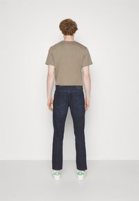 T-shirt marrone a maniche corte, jeans blu scuro e sneaker bianche con dettagli verdi, viste da dietro su uno sfondo bianco semplice.