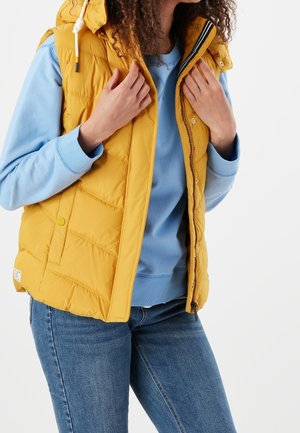 Person iført en gul quiltet vest over en lyseblå sweatshirt og blå jeans, der justerer vestens krave med begge hænder.
