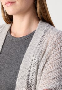 Lichtgrijze gebreide cardigan met een open voorkant, voorzien van een losse, luchtige textuur en brede mouwen, gedragen over een donkergrijze getailleerde shirt.