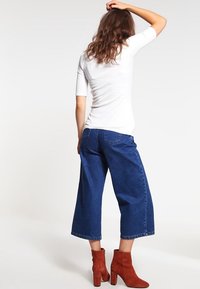 Frau mit langen braunen Haaren, die ein weißes kurzärmeliges Oberteil, blaue Jeans mit weitem Bein und rostfarbene Stiefeletten mit Absatz trägt, steht mit der Hand am Kopf.