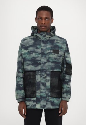 Multi-toned green and blue patterned jacket with a hood, featuring mesh side pockets and a solid black patch on the chest. Textured fabric.