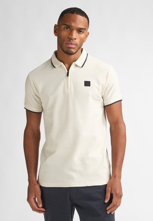 Polo crème avec un motif texturé, col zippé, accents bleu marine sur le col et les manches, et un petit logo carré foncé sur la poitrine.