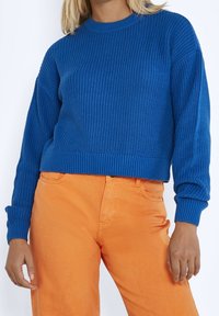 Blå stickad tröja med ribbdesign och långa ärmar, parat med orangea byxor. Tröjan har rund halsringning och kort skärning.