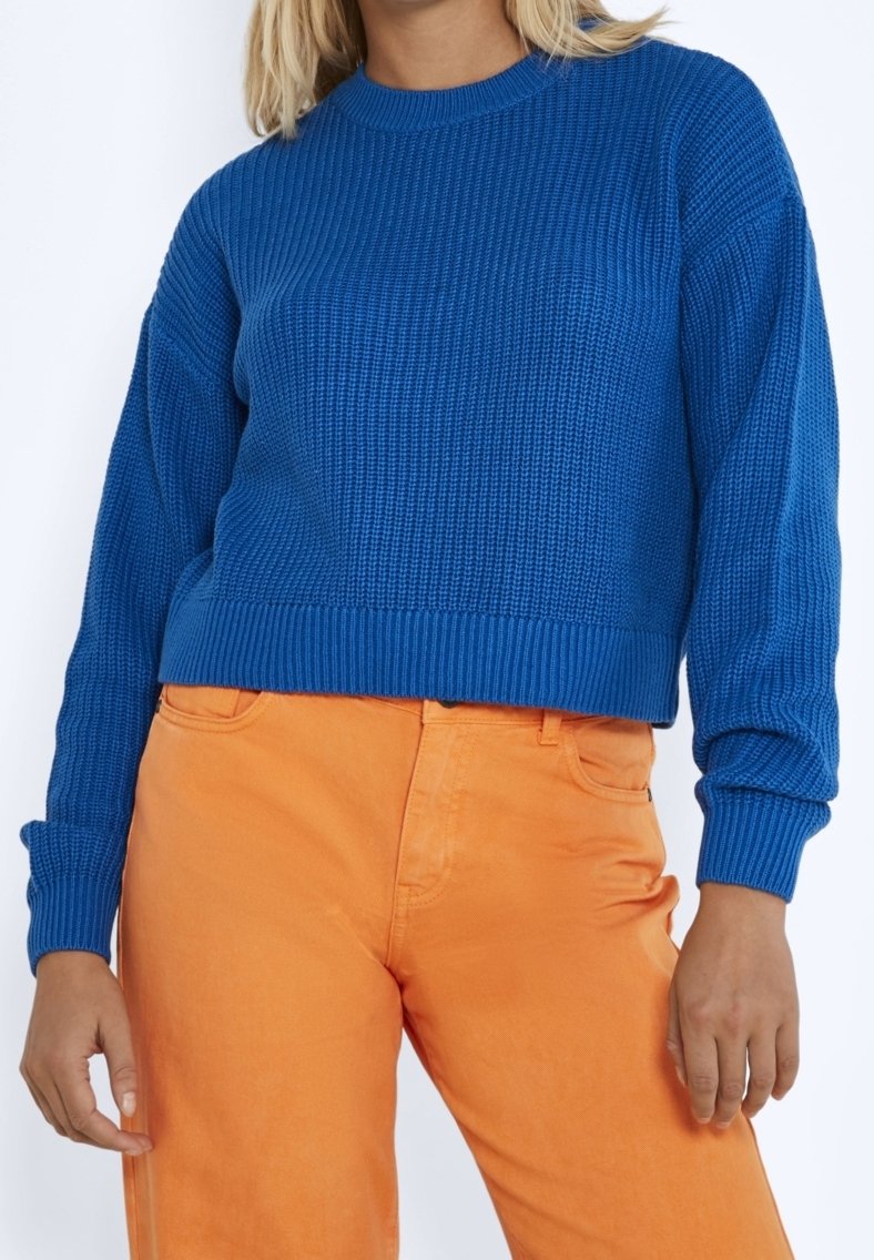 Blå stickad tröja med ribbdesign och långa ärmar, parat med orangea byxor. Tröjan har rund halsringning och kort skärning.