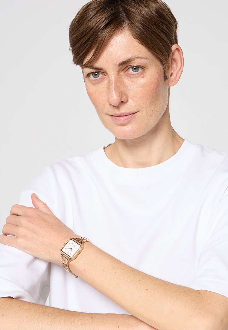 Frau mit braunen kurzen Haaren, die ein weißes Hemd und eine rechteckige goldene Armbanduhr am linken Handgelenk trägt, vor einem neutralen Hintergrund.