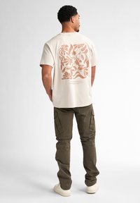 Beige katoenen t-shirt met een bruine bloegraphic op de achterkant, gecombineerd met olijfgroene cargobroeken en witte sneakers.