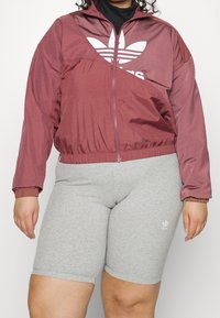 Vínově červená větrovka na zip s elastickým pasem a manžetami, zdobená bílým logem Adidas; doplněná světle šedými elastickými cyklistickými šortkami.