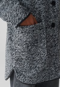 Abrigo gris texturizado con acentos negros, que presenta un bolsillo visible, dobladillo asimétrico y botones para un cierre seguro. La tela es suave y gruesa.