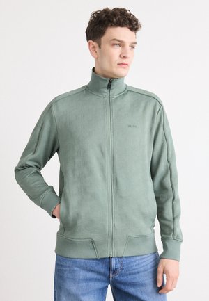 Lys grønn zip-up hettegenser med et teksturert geometrisk mønster, ribbede mansjetter og krage, kombinert med blå jeans.