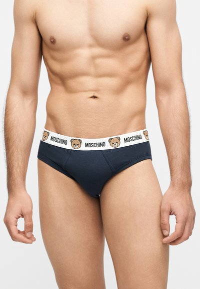 Ανδρικός κορμός με ναυτικό μπλε εσώρουχα με λευκή μέση, διακοσμημένη με καφέ πρόσωπα αρκουδάκια και επαναλαμβανόμενο κείμενο "MOSCHINO".