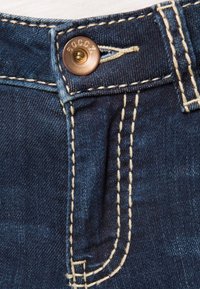 Los jeans de mezclilla azul oscuro presentan un cierre de botón de bronce, costuras amarillas en contraste y un pequeño bolsillo frontal. La textura parece suave.